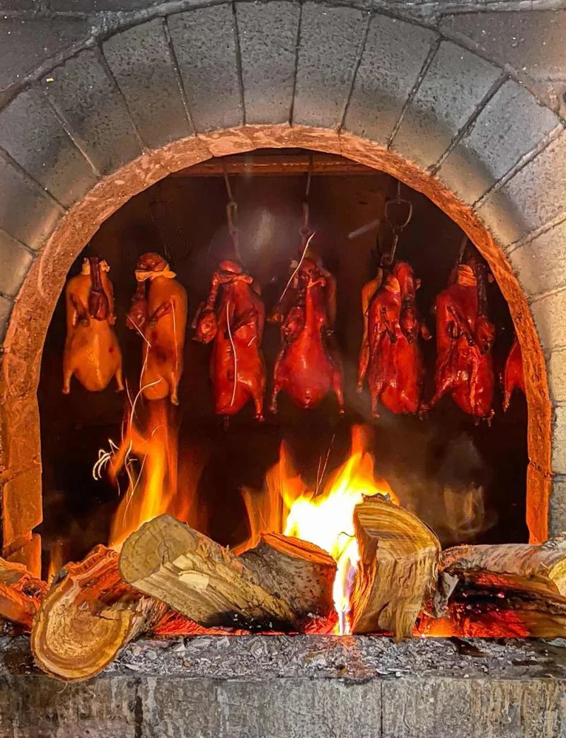 Whole Peking Ducks roasting inside a traditional brick oven over an open flame.