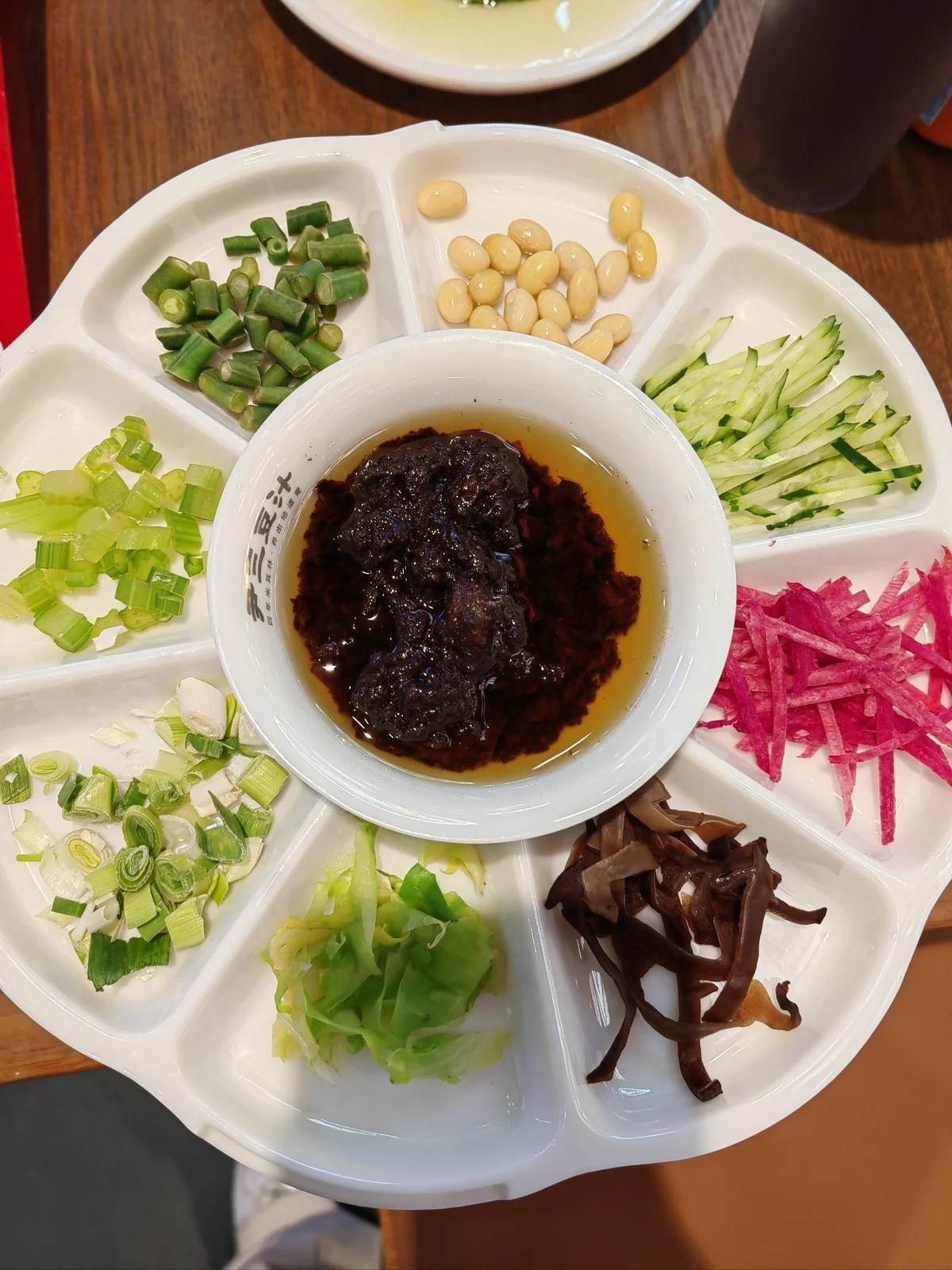 A platter of Zhajiang Noodle toppings featuring cucumber strips, radish strips, soybeans, wood ear mushrooms, cabbage, and scallions with Zhajiang sauce in the center.