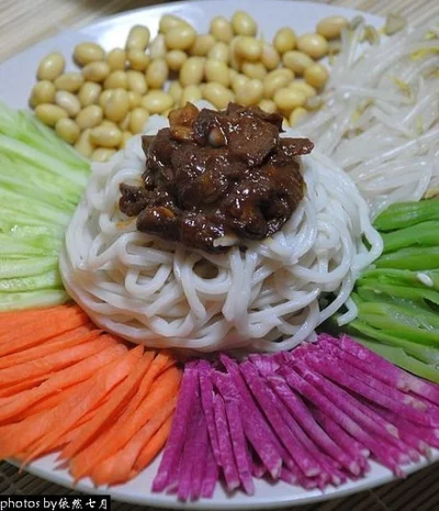 A bowl of noodles topped with Zhajiang sauce surrounded by colorful fresh toppings including soybeans, bean sprouts, cucumber, green beans, radish, and carrots.