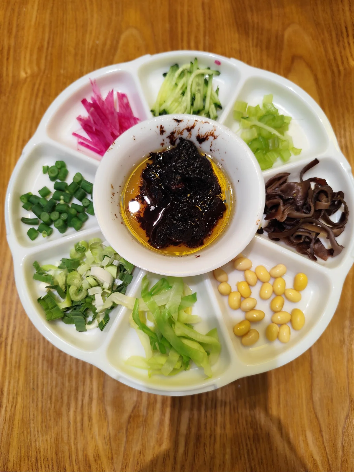 Freshly sliced Zhajiang Noodle toppings displayed in small sections, including cucumber, radish, green onions, celery, soybeans, and wood ear mushrooms.