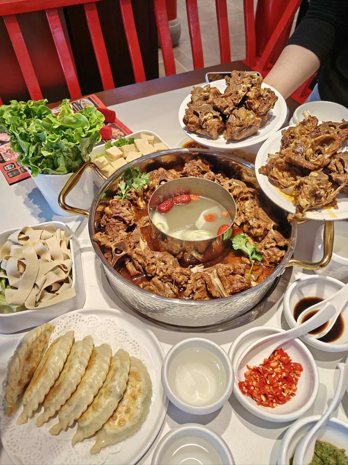 Large family-style lamb spine hotpot feast with simmering marrow-rich lamb bones surrounded by vegetables, tofu, dumplings, and dipping sauces, showcasing Beijing’s communal dining culture.