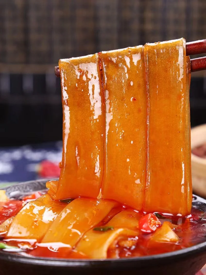 Wide kuanfen noodles coated in spicy red broth, lifted by chopsticks to show their glossy texture and smooth, chewy surface.