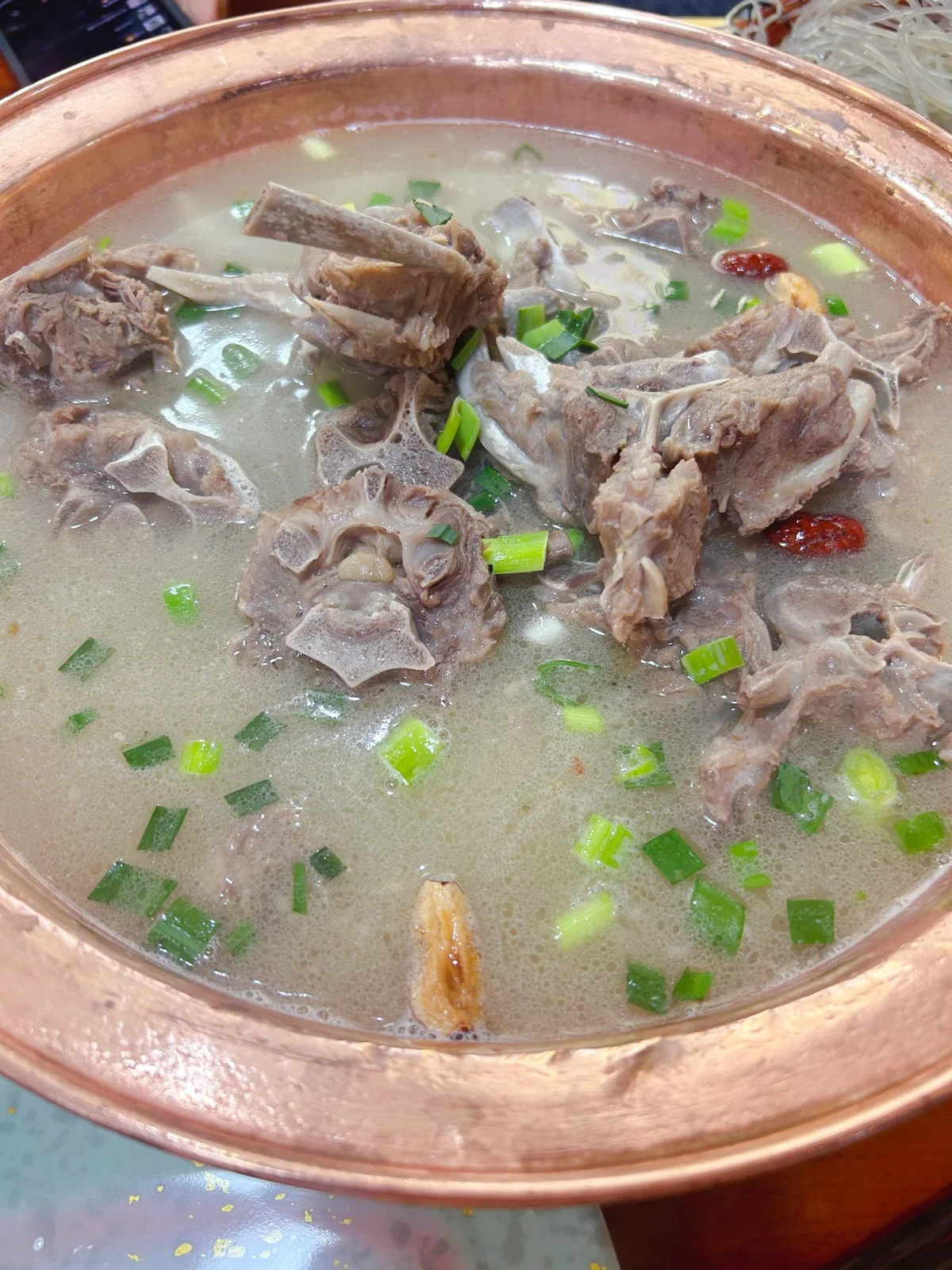 A copper pot filled with clear-simmered lamb spine broth, with visible goji berries and scallions floating on the surface.
