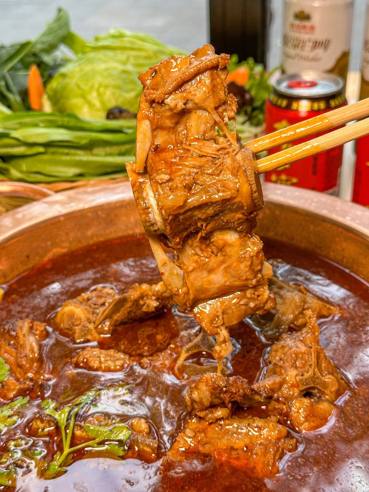 Chopsticks lifting a tender piece of lamb spine from a rich, simmering Yang Xiezi hotpot, highlighting the marrow-filled bone and deep red broth.
