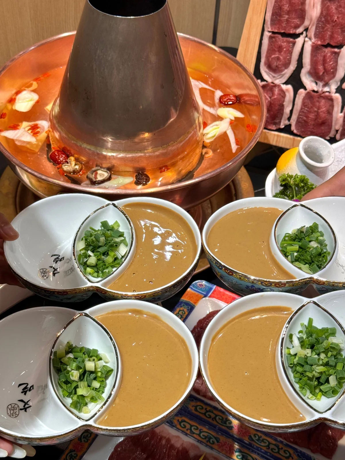 Bowls of sesame dipping sauce garnished with chopped scallions beside a copper hotpot simmering with clear broth and aromatics for instant-boiled mutton.