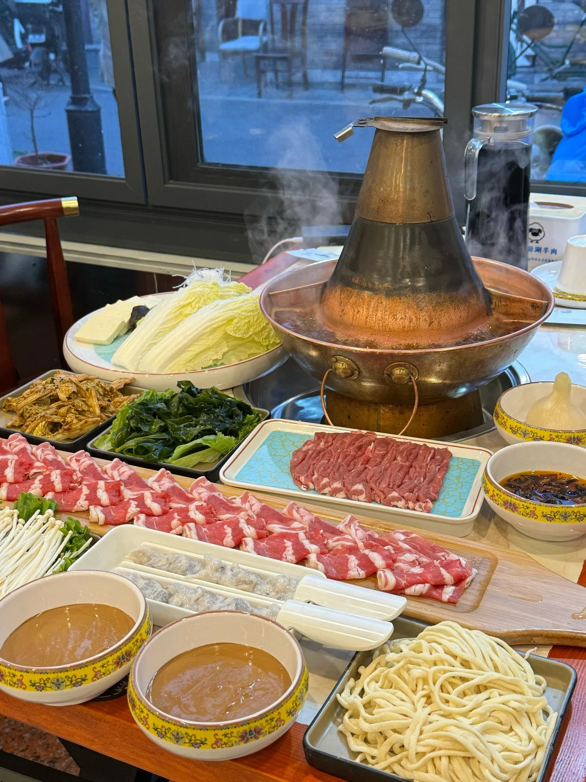 Traditional Beijing copper hotpot boiling with clear broth, surrounded by thinly sliced lamb, fresh vegetables, tofu, and sesame dipping sauce for authentic instant-boiled mutton.