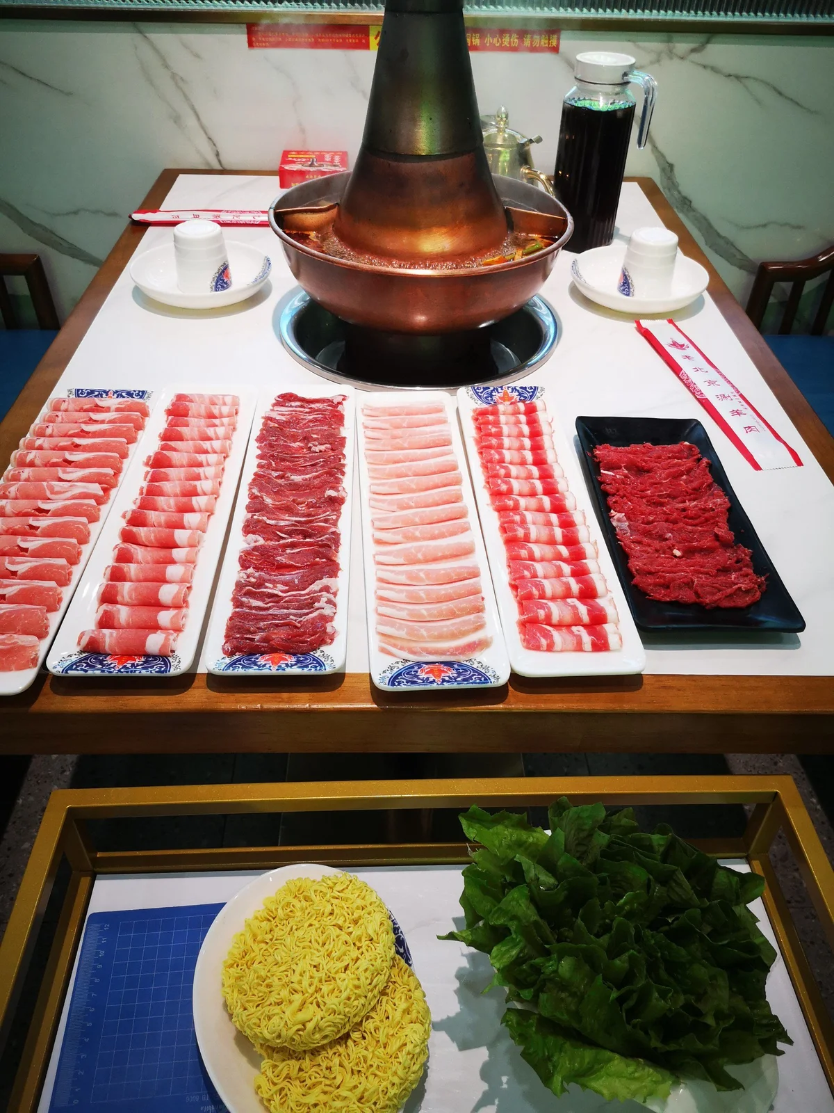 Neatly arranged lamb slices, leafy greens, and noodles displayed beside a boiling copper hotpot prepared for traditional Beijing instant-boiled mutton.