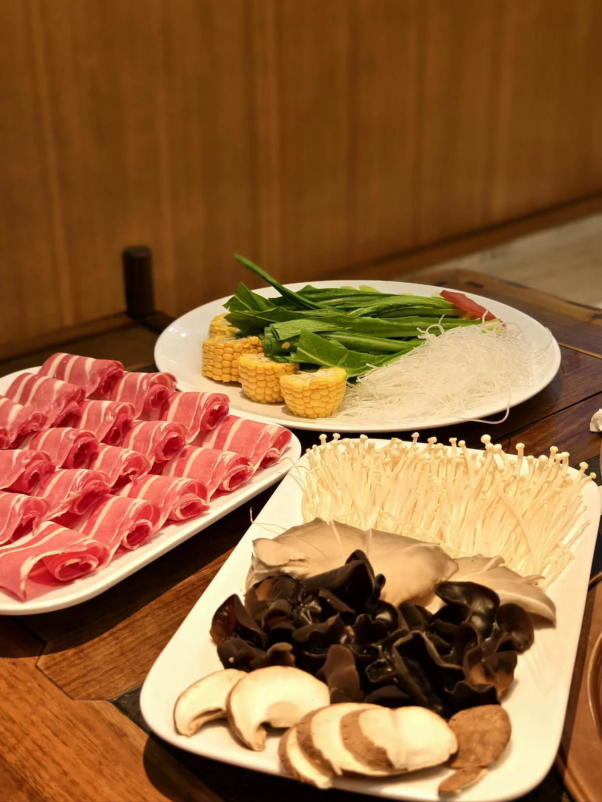 Fresh ingredients for Beijing instant-boiled mutton, including thinly sliced lamb, assorted mushrooms, enoki, black fungus, corn, leafy greens, and vermicelli arranged on serving plates.