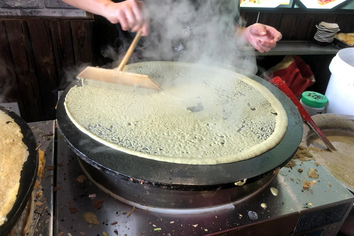 Jianbing batter being spread thinly across a round griddle