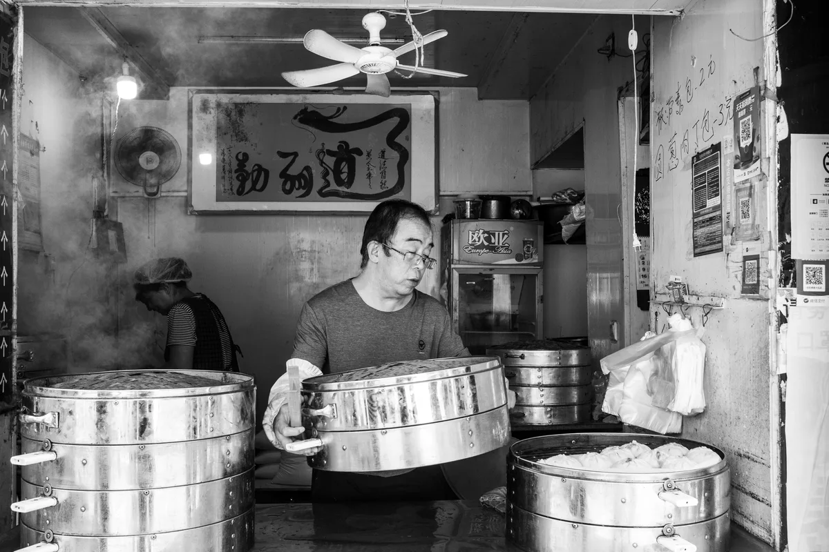 Baozi shop worker handling large steamers with buns ready for breakfast service