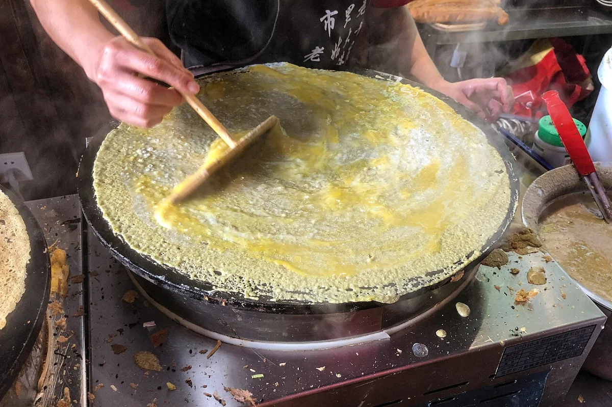 Jianbing batter being spread thinly across a round griddle before egg and fillings are added