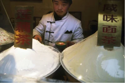 A Beijing snack-counter cook preparing savoury miancha beside bowls and sesame ingredients